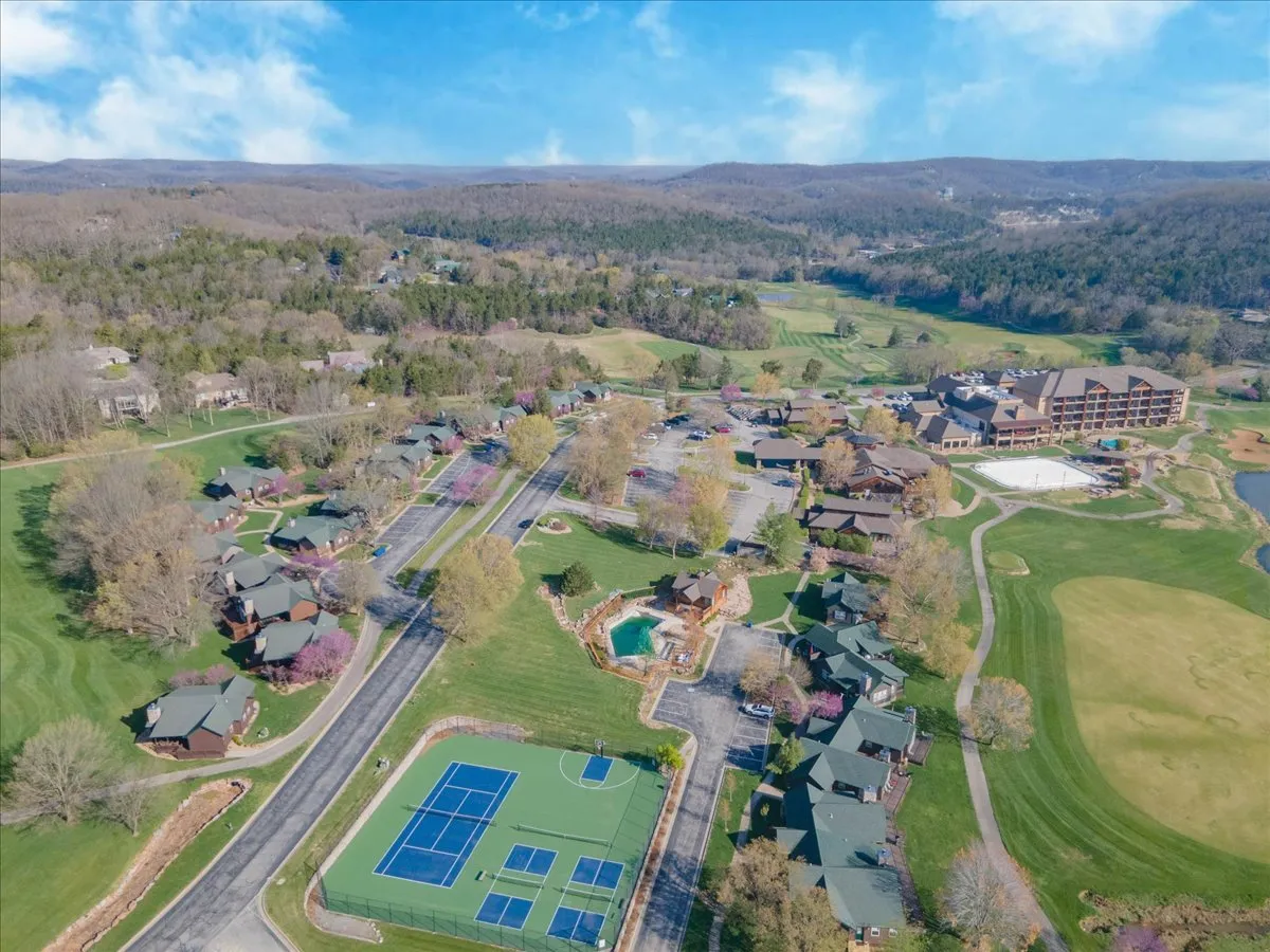 A wide view of the resort grounds featuring tennis courts, a swimming pool, a lodge, and surrounding natural beauty. The scene is lively yet tranquil, offering multiple options for recreational activities set against a backdrop of trees and greenery.
