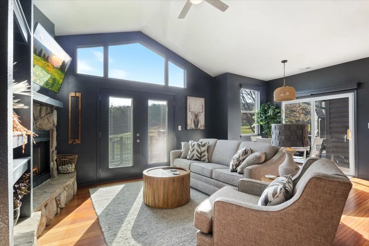 A spacious living room featuring large windows that flood the space with natural light and provide sweeping views of the outdoors, possibly a golf course. The room is anchored by a cozy stone fireplace, comfortable seating including a plush sofa and armchairs, a wooden coffee table, and a wall-mounted TV. Modern and inviting décor completes the look.