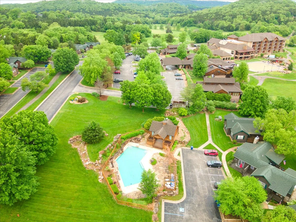 A vibrant community swimming pool surrounded by green landscaping and wooden pavilions, with nearby cabins and a parking lot visible in the background. The scene conveys active family fun and a strong connection to nature.
