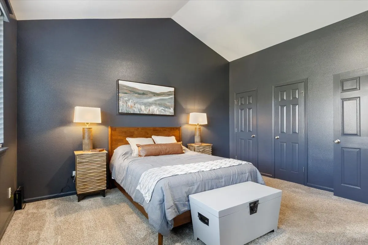 A tranquil bedroom setting with a neatly made bed, wall art above, nightstands with lamps on either side, and a chest for storage at the foot of the bed. The mood is peaceful and restful, ideal for ending or starting the day.