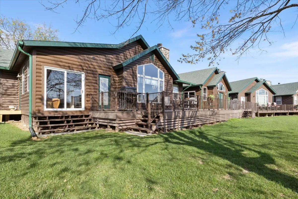 The exterior of a cozy log cabin, featuring large windows and a spacious deck area ideal for outdoor gatherings. The setting invites guests to imagine relaxing, grilling, or sharing a meal as the sun sets over the peaceful natural surroundings.