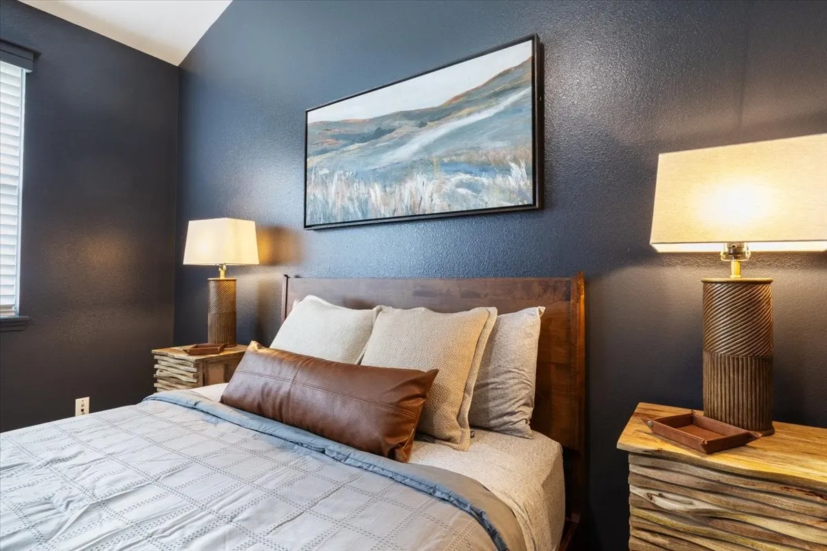 A cozy bedroom scene focusing on a bed layered with plush pillows and soft linens, accompanied by tasteful decorative artwork and matching table lamps on each side. The styling feels inviting and geared toward comfort and relaxation.
