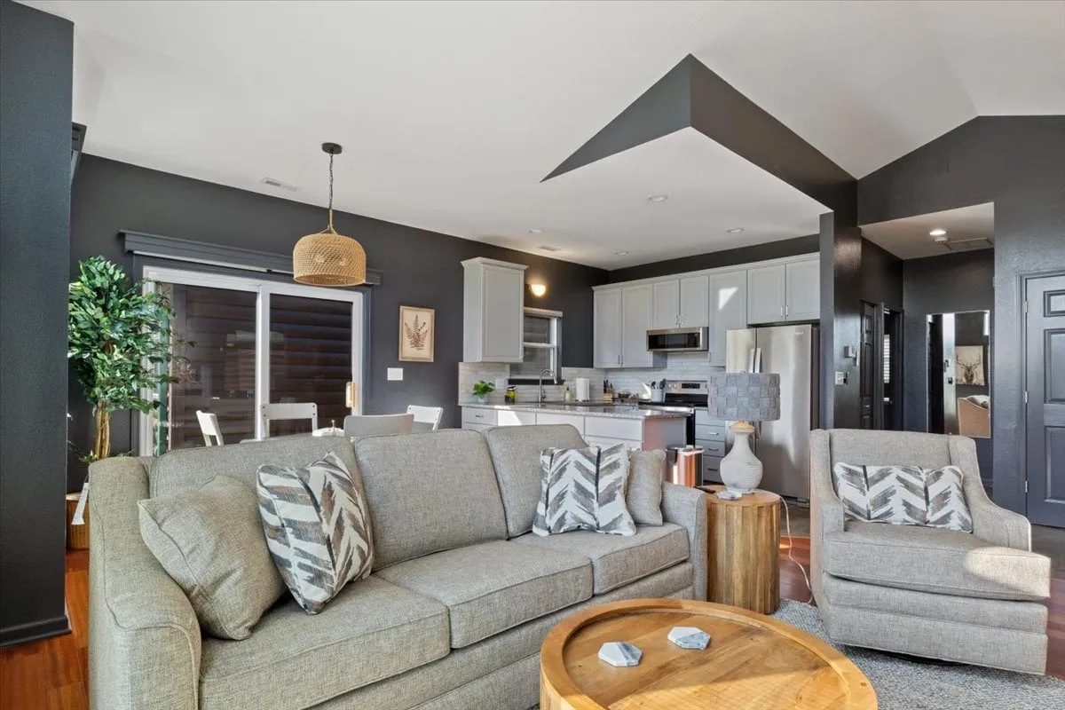 An open-concept living area with a contemporary sofa, coffee table, kitchen area in the background, decorative plant, and statement wall art. The light, neutral palette and modern finishes create a comfortable, inviting setting suitable for both relaxing and entertaining guests.