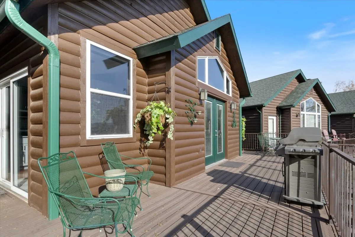 A charming log cabin exterior with green accents, a welcoming deck furnished with seating, and planters adding pops of color. The scene is set amidst lush greenery, evoking a cozy, private haven perfect for couples seeking a romantic escape. The deck suggests a spot for morning coffee or sunset drinks while enjoying nature.