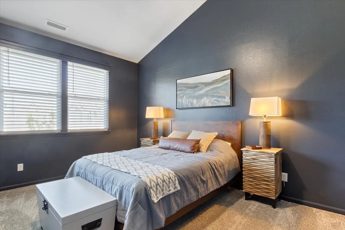 A sunlit bedroom featuring a queen-sized bed adorned with decorative pillows and soft linens, flanked by modern bedside lamps and complemented by framed artwork on the wall. Windows allow natural light to gently filter into the space, creating a serene and cozy atmosphere.